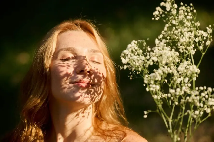 Image of a woman with her eyes closed, basking in the sun with flowers casting shadows on her face.