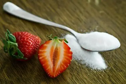 Image of a strawberry sliced in half next to a spoon overflowing with an artificial sweetener like aspartame.