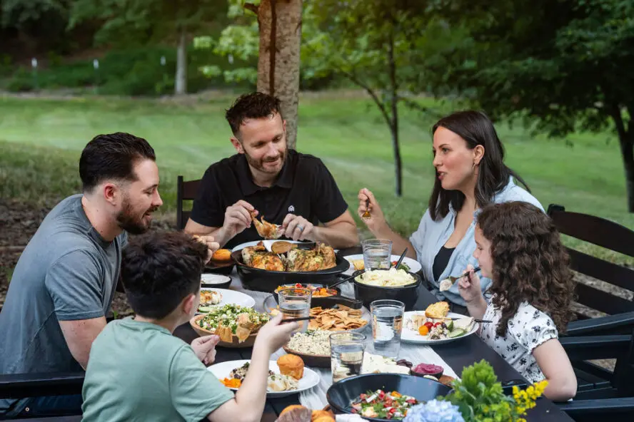 Image of a family sitting at a table outside enjoying a summer dinner using the Xtrema Founder's Classic Collection cookware pieces.