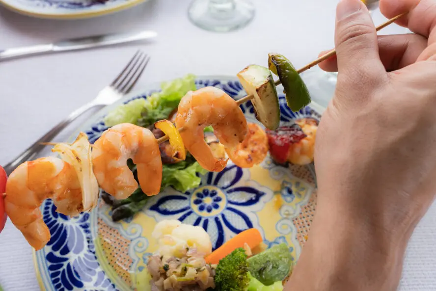 Image of a hand holding a shrimp and vegetable skewer over a plate.