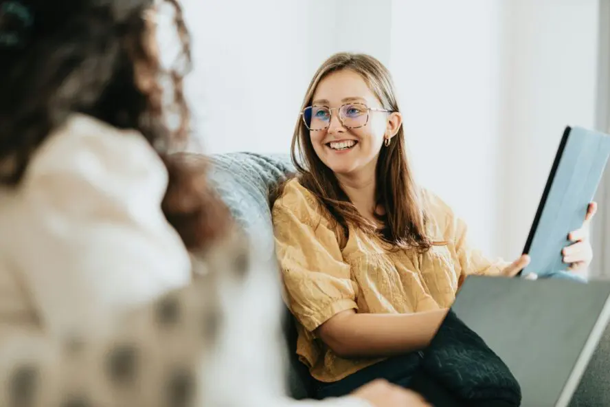 Image of a young woman with glasses smiling at another woman in conversation sitting on a couch.