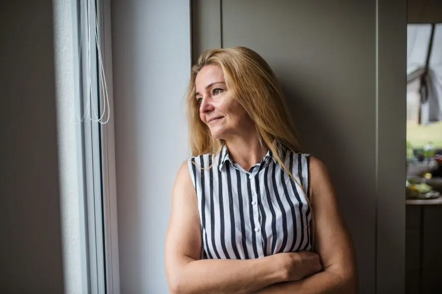 A blonde middle-aged woman standing with her arms crossed, looking out a window with a small smile on her face.