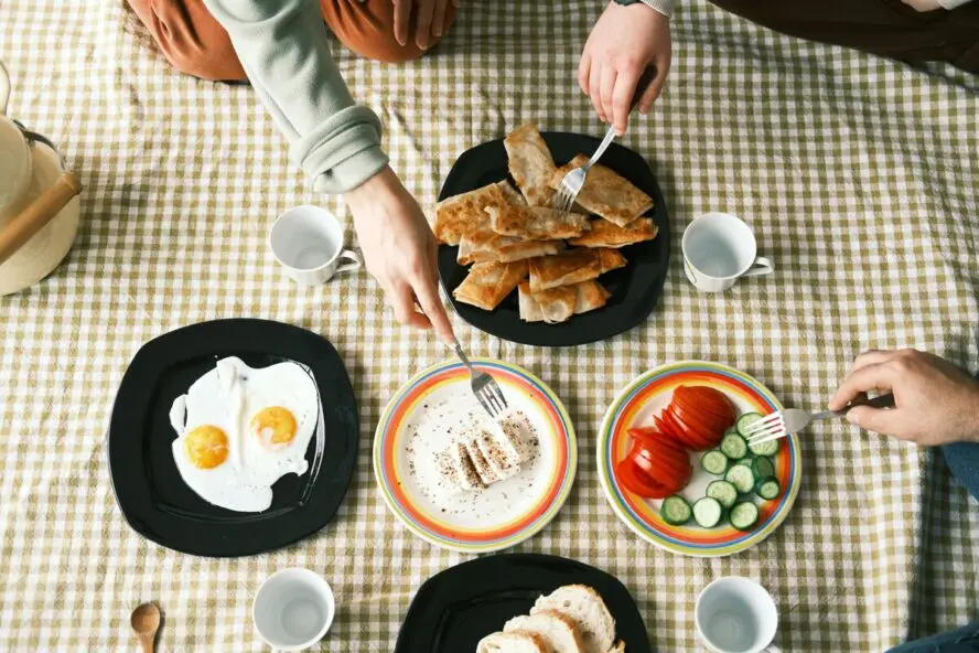Image of various breakfast dishes spread out on a checkered tablecloth, from a couple of fried eggs to a spread of cut cucumbers and tomatoes.