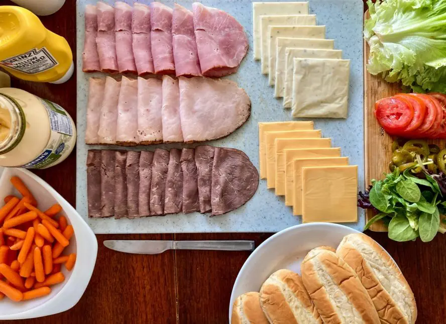 Image of a spread of deli meat cold cuts, various sliced cheeses, and other sandwich ingredients laid out on a board.