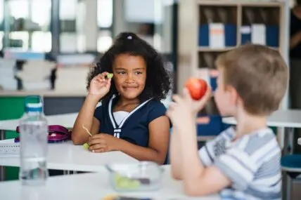 Image of two schoolchildren sitting at schooldesks and comparing snacks — an apple and grapes.