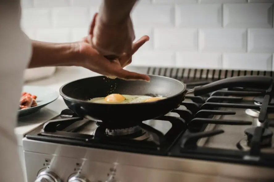 Image of a nonstick pan on the stovetop frying eggs, with a pair of hands grinding salt over them.
