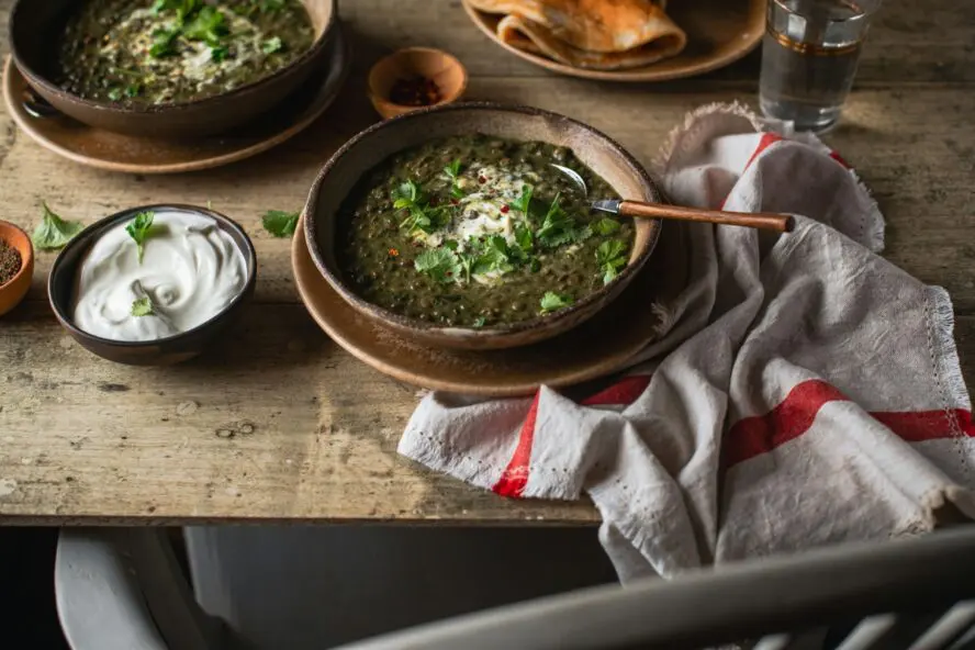 Image of a bowl of rustic lentil soup, topped with fresh green onions and served on a wooden table alongside a dish of sour cream.
