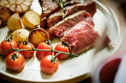 Image of a plate with slices of medium rare steak, roasted potatoes, and cherry tomatoes on the vine.