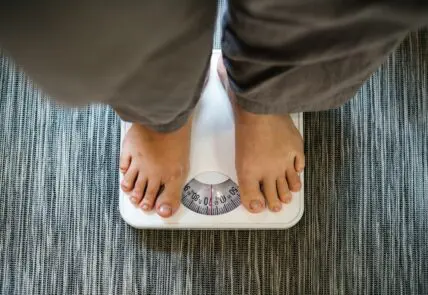 Image of a scale shot from above, showing the legs and feet of a woman wearing green pants.