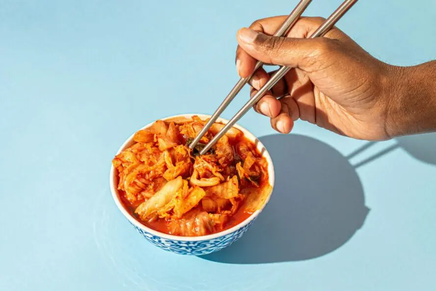 Image of a white bowl full of kimchi and a hand digging in with chopsticks, all set on a light blue background.