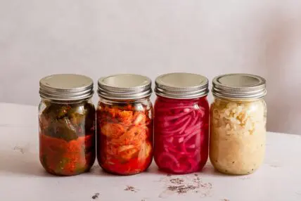Image of four mason jars lined up in a row and filled with colorful fermented foods like kimchi and sauerkraut.
