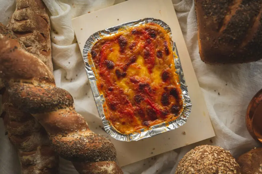Image of a gooey, cheesy tin of lasagna displayed on a table surrounded by loaves of bread.