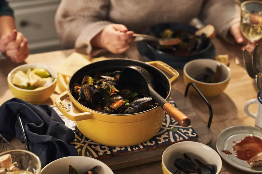 Image of a round Staub Dutch oven in the colorway Citron, shown on a dining table with a stew of mussels and a large serving spoon.