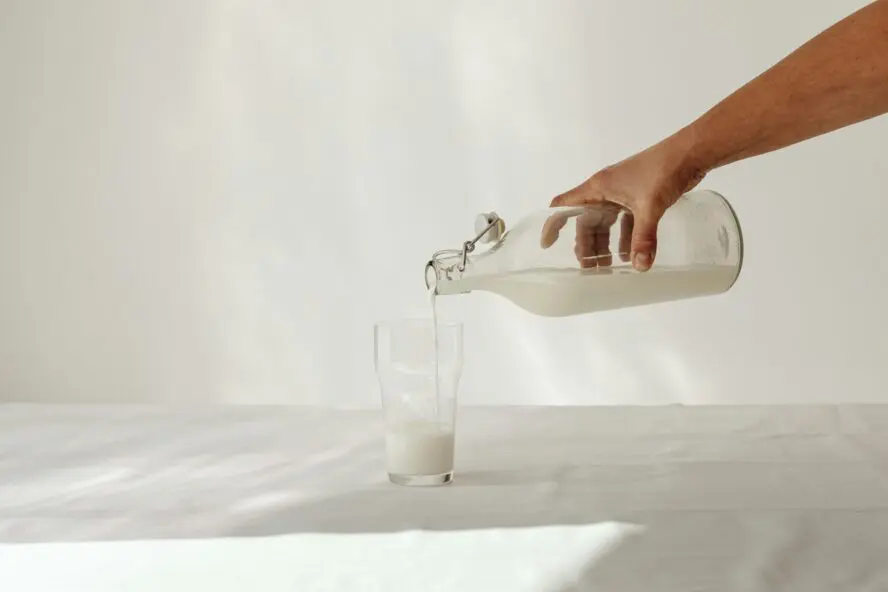 Image of a hand pouring milk from a glass bottle into a glass, all against a white background.