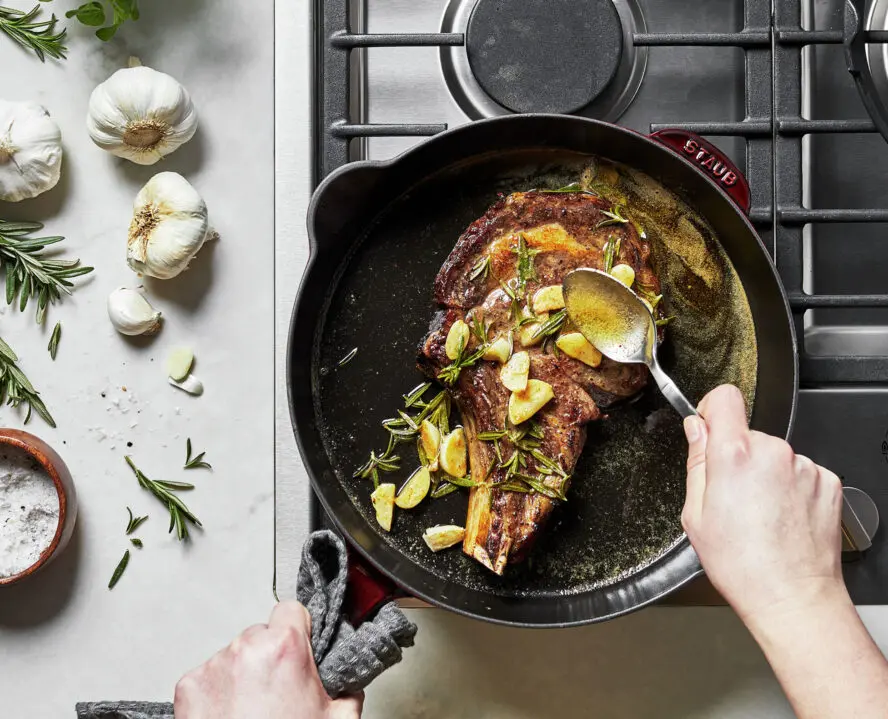 Image of a Staub Traditional Deep Skillet in Grenadine on a stovetop, with a pair of hands using a spoon to baste a steak with butter, topped with rosemary and garlic.