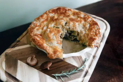 Image of a gorgeously buttery chicken pot pie in a dish with a slice taken out and displayed on a wooden board.