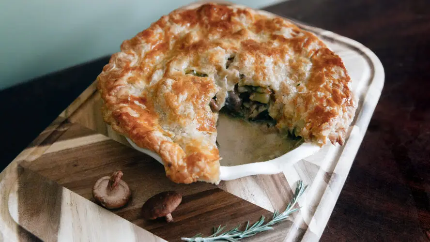 Image of a gorgeously buttery chicken pot pie in a dish with a slice taken out and displayed on a wooden board.