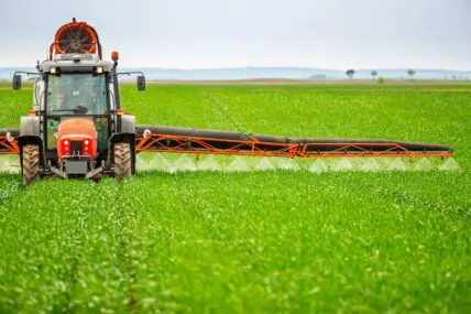 Image of a large tractor driving over a field of crops, spraying herbicides and pesticides as it goes.