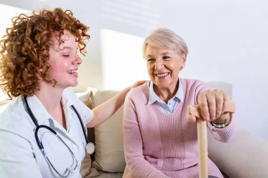 Image of an elderly woman with a cane sitting on a couch next to a nurse with a stethoscope around her neck.