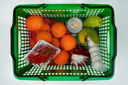 Image of a green grocery basket filled with produce items like oranges, garlic, cabbage, and chilis.