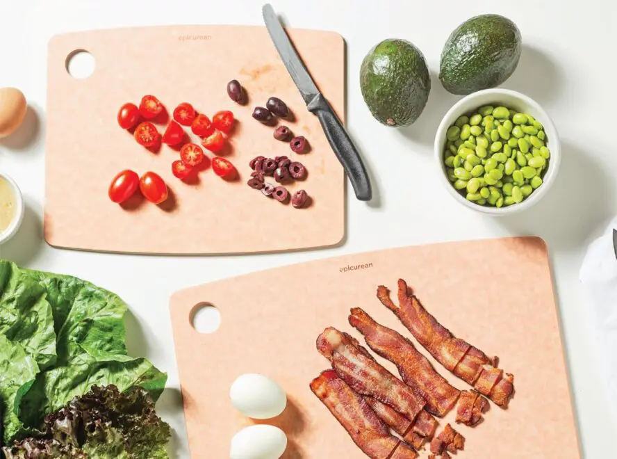 Image of two Epicurean composite cutting boards, one being used to chop olives and cherry tomatoes and the other slicing bacon. 