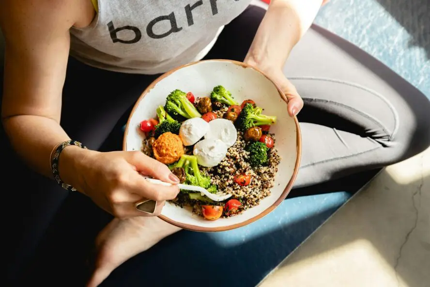 Image of a woman sitting cross-legged in workout clothing holding a grain bowl with broccoli, tomatoes, and mozzarella over her lap.