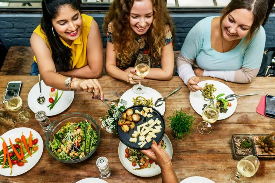 Image of several women sitting around a dining table and sharing plates of food and glasses of wine.