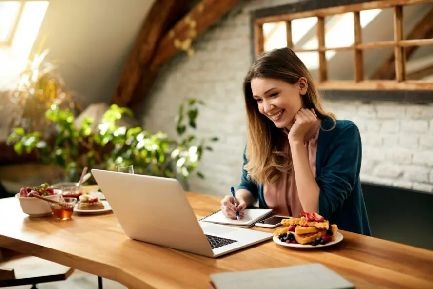 Image of a woman smiling at her laptop while taking notes with a plate of waffles beside her.