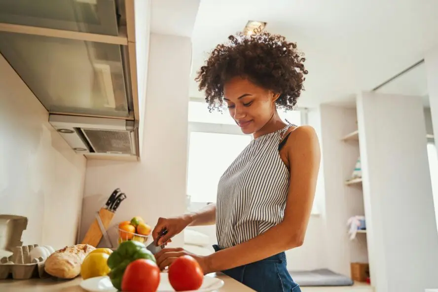 Image of a young woman in a kitchen standing at the counter prepping vegetables on a cutting board in front of her.