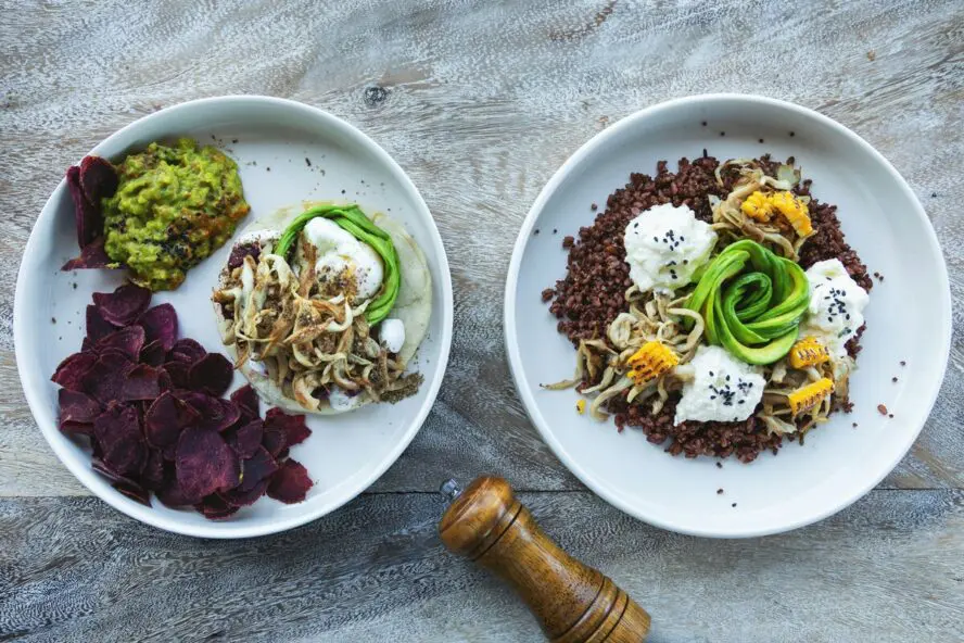 Two plates with colorful dishes featuring avocado, beetroot chips, grains, corn, and sprouts.