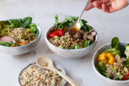 Image of several grain bowls with various vegetables, one of which is being topped with dressing from a spoon.