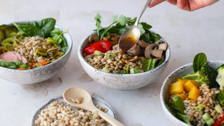 Image of several grain bowls with various vegetables, one of which is being topped with dressing from a spoon.
