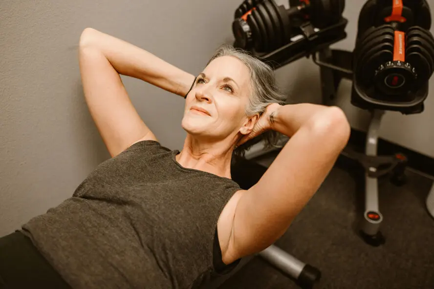Image of an older woman laying on a workout bench with her hands behind her head as she does a sit-up.