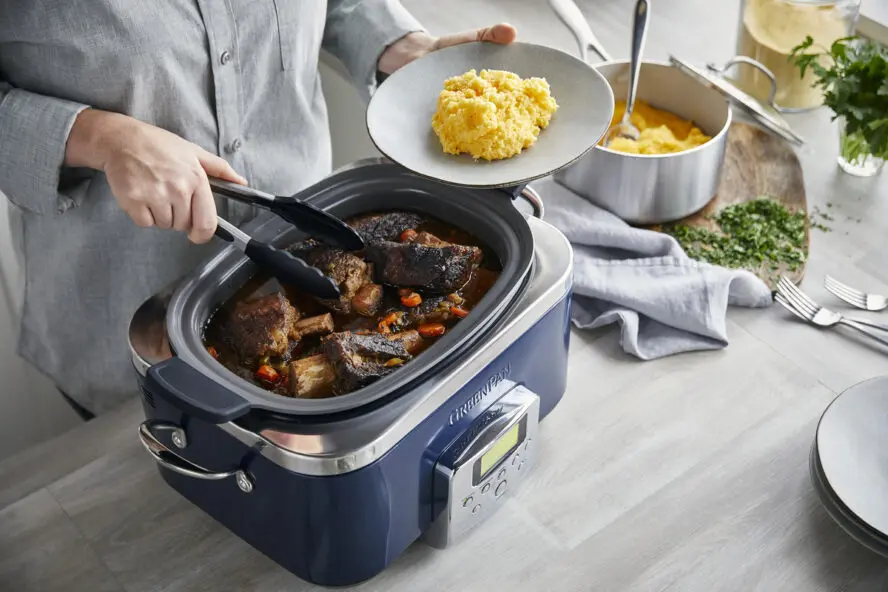 Image of the GreenPan Elite Slow Cooker in Oxford Blue, shown with a pair of hands removing short ribs and plating them on a bed of grits.