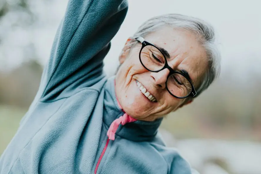 Image of an older woman wearing glasses and stretching with her arm over her head and her eyes closed.