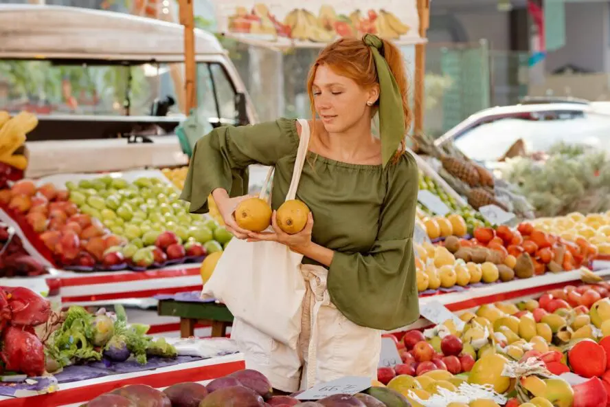 Image of a young redhead woman shopping at a colorful outdoor market, putting fruit into a reusable tote bag.