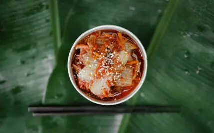 Image of a small bowl of kimchi alongside a pair of chopsticks, displayed on a large banana leaf.