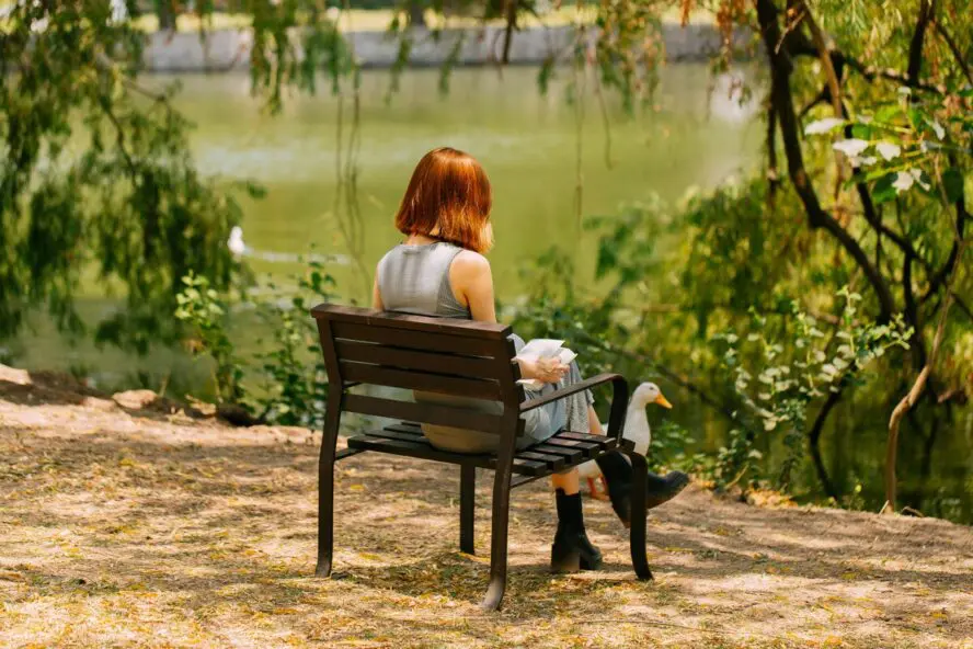 Image of a young woman with red hair sitting on a park bench by a tranquil pond.