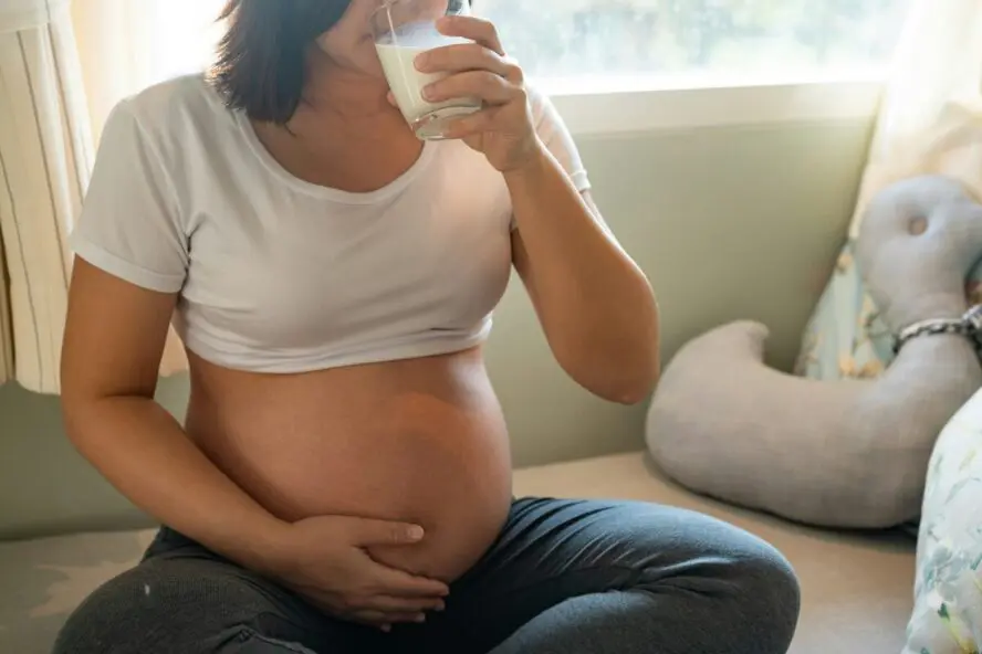 Image of a pregnant woman holding her stomach and sitting cross-legged on a couch as she drinks milk.