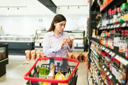 Image of a young woman at a supermarket pushing a cart and examining the ingredients on a can of food.