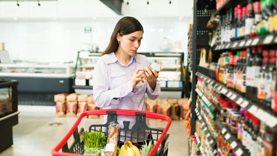 Image of a young woman at a supermarket pushing a cart and examining the ingredients on a can of food.