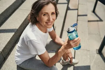 Image of a middle-aged woman holding a water bottle and smiling at the camera from where she sits on outdoor steps.