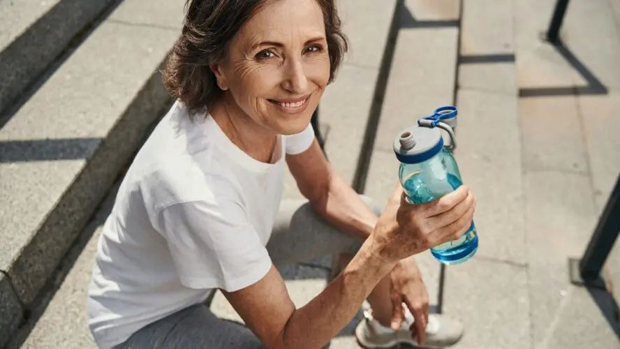 Image of a middle-aged woman holding a water bottle and smiling at the camera from where she sits on outdoor steps.