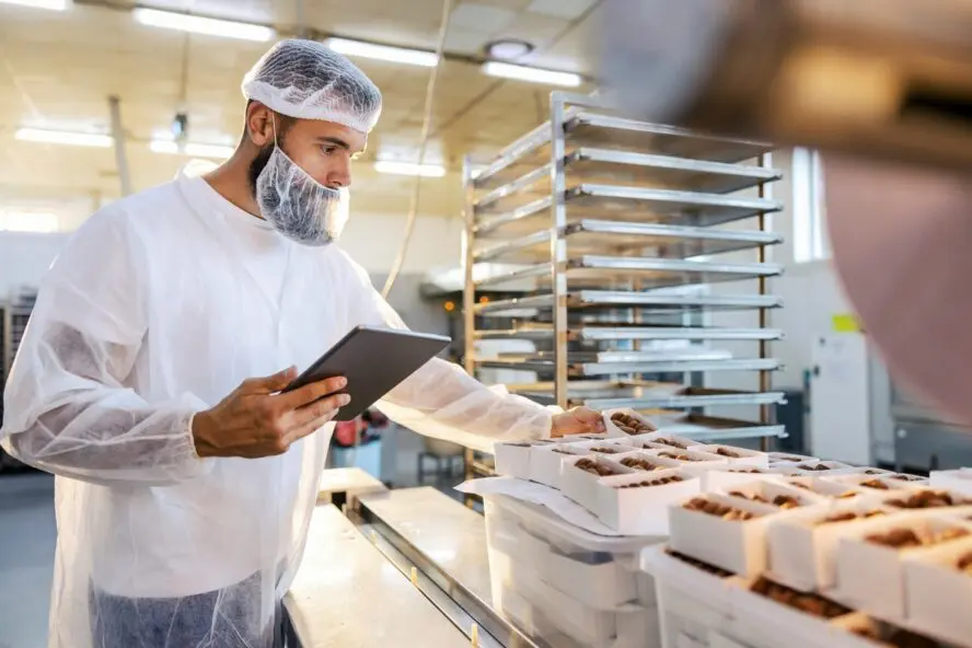 Image of a man wearing a mask, hairnet, and lab coat inspecting food items in a factory with an iPad in hand.