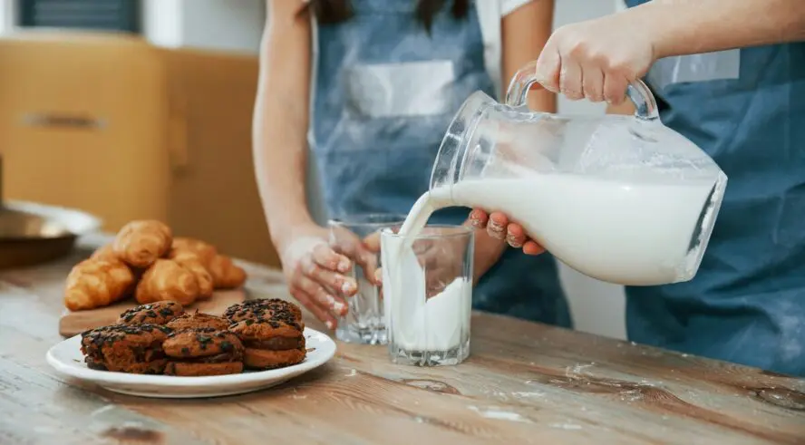 Image of two kids pouring a glass of milk from a jug to enjoy with a plate of pastries.