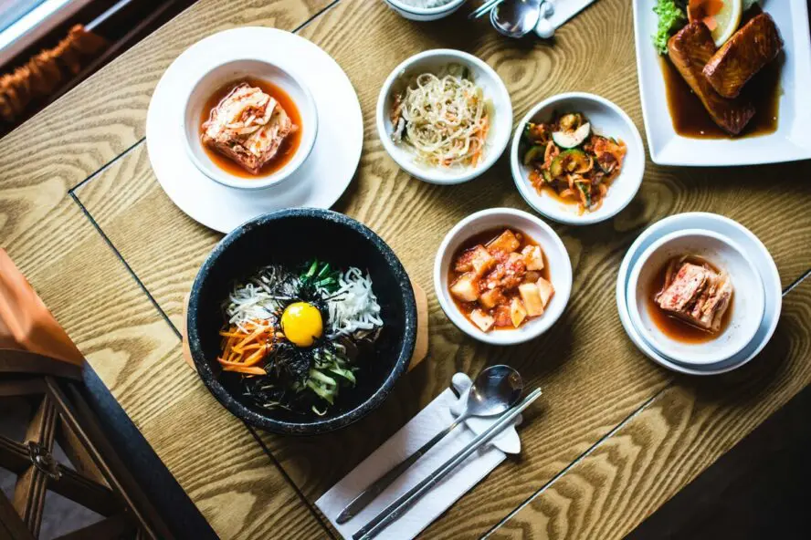 Image of a spread of dishes displayed on a wooden table with bibimbap, several small kimchi side dishes, and table settings.