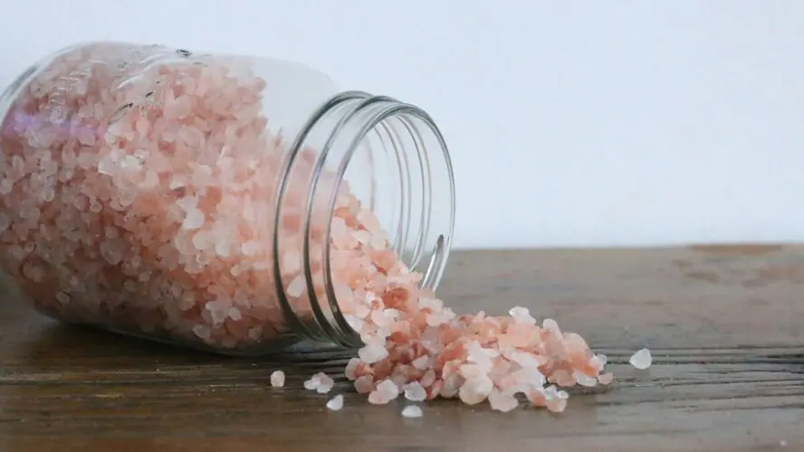 Image of a glass mason jar of pink Himalayan salt spilling out onto a wooden table.
