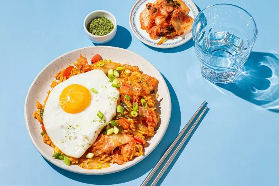 Image of a plate of kimchi fried rice (with a side dish of kimchi and a glass of water) topped with a fried egg and set on a light blue background.