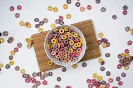Image of a white bowl of cereal on a wooden board, with cereal spilling all over the surroundings.