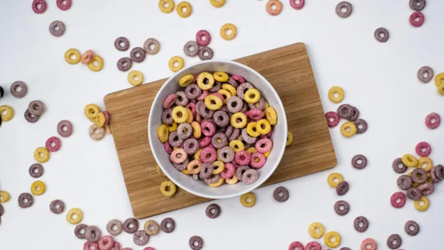 Image of a white bowl of cereal on a wooden board, with cereal spilling all over the surroundings.